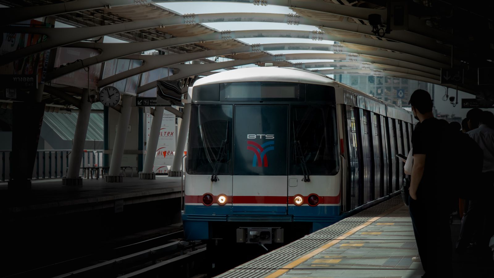 A BTS Skytrain pulls into a covered elevated platform in Bangkok as passengers wait behind the yellow line