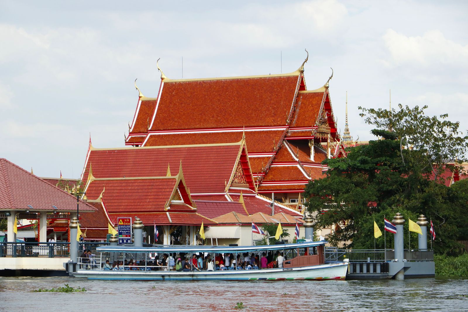 A covered passenger boat full of riders sits at a pier on the Chao Phraya River in front of a temple with red and gold roofs