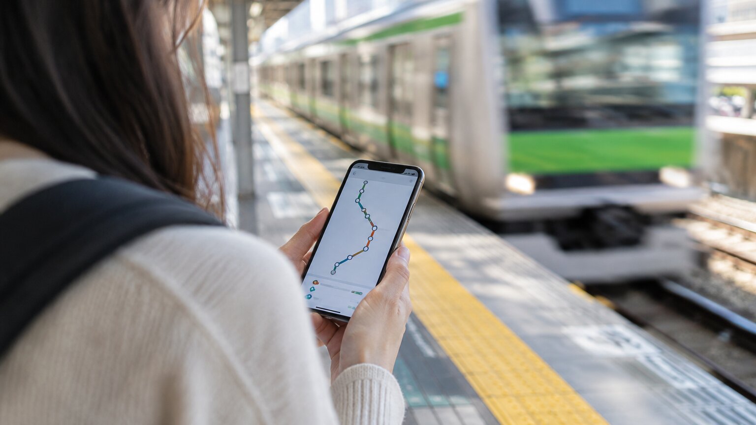 A traveler on a train platform in Japan checks a route on a transit app while a green-striped train pulls in