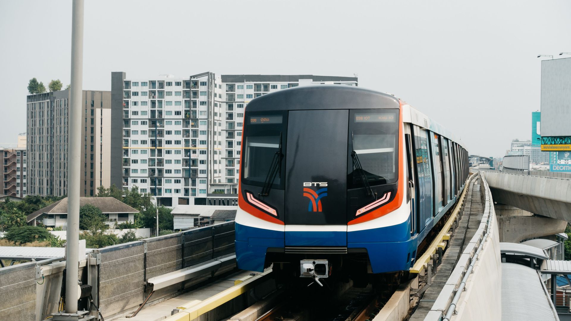 A blue, white and orange BTS Skytrain approaches on an elevated track in Bangkok, with apartment blocks behind it