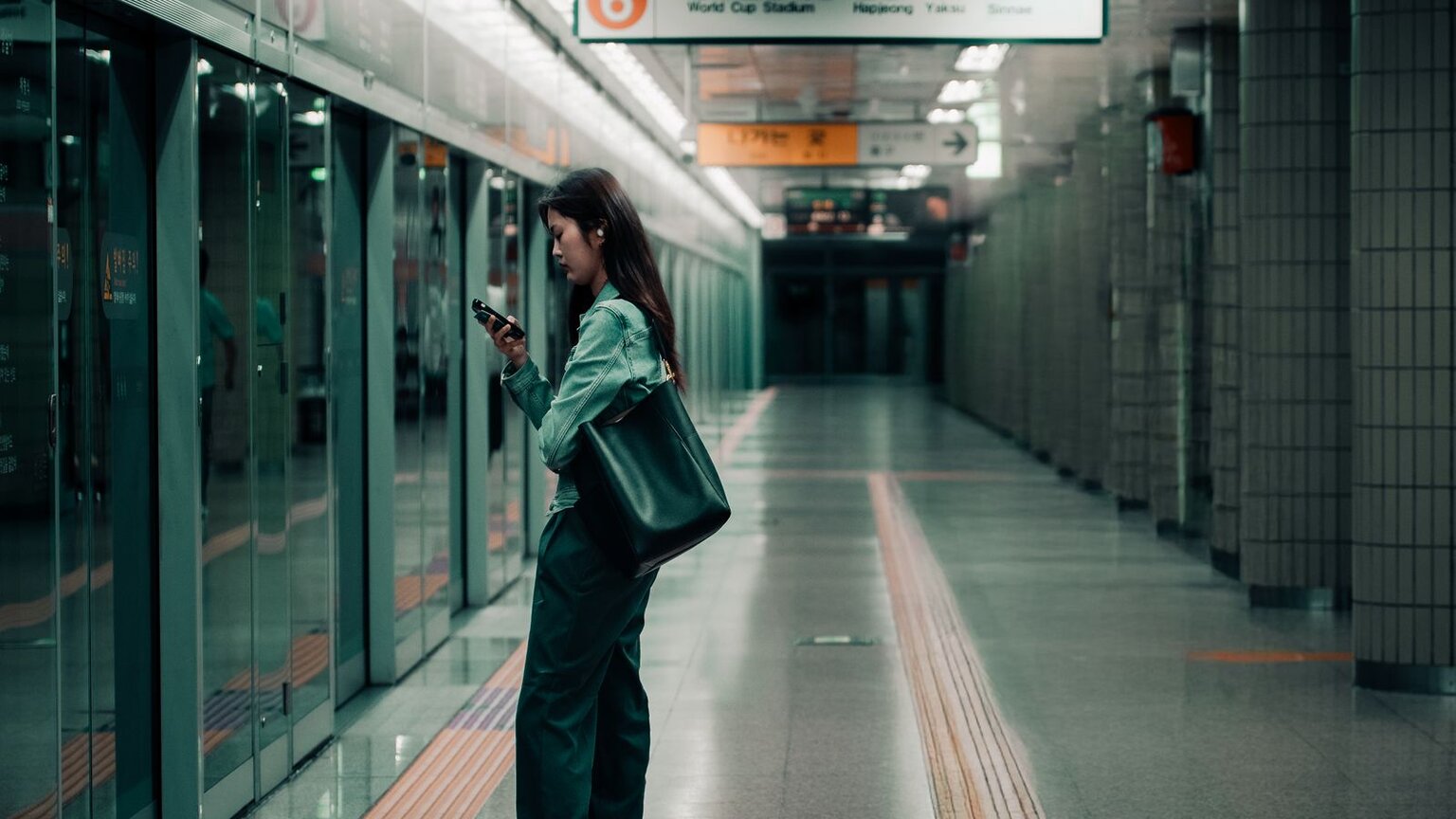 A woman looks down at her phone on a Seoul subway platform, with platform screen doors on the left and Korean and English signs overhead