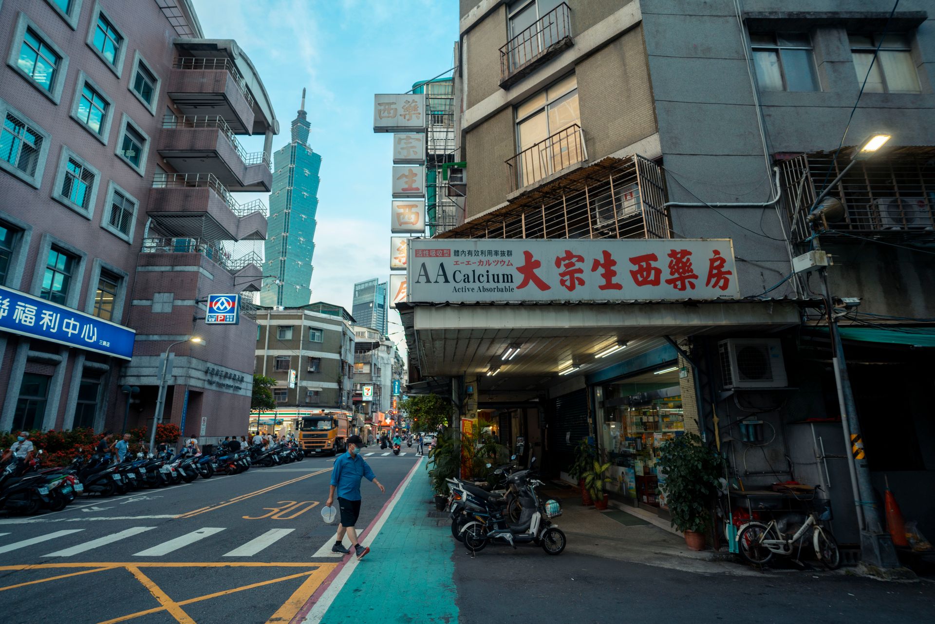A man crosses a quiet Taipei side street lined with parked scooters and a pharmacy sign, with Taipei 101 rising between the buildings behind him