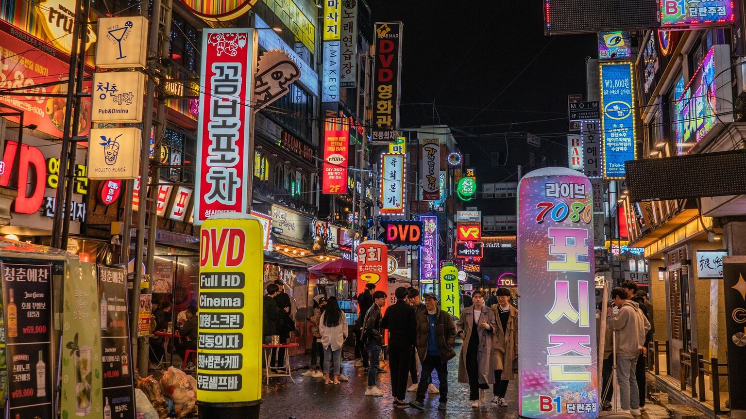 A Seoul street at night lined with Korean neon signs, with people walking between the shops