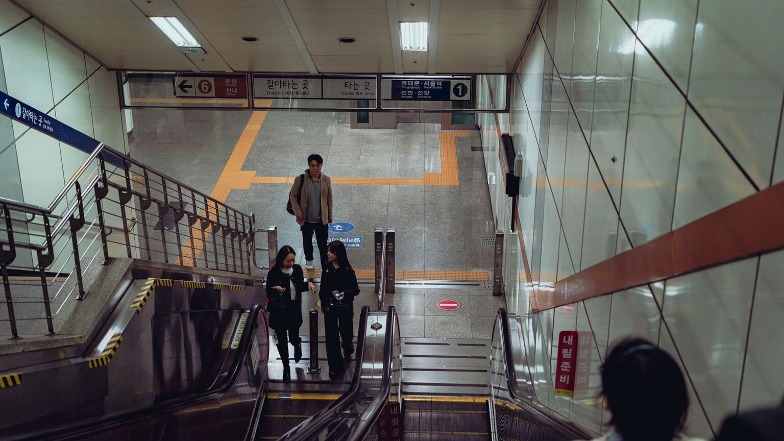A transfer corridor inside a Korean subway station, with passengers walking past the escalators under multilingual transfer and platform signs
