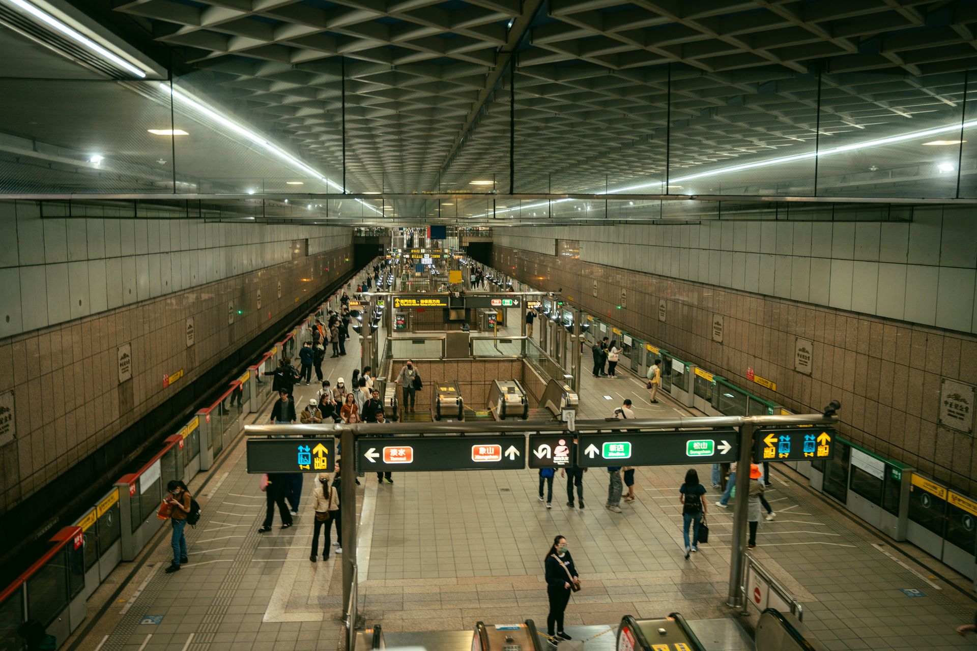 Passengers walk along a long underground Taipei Metro platform beneath bilingual direction signs and a coffered ceiling