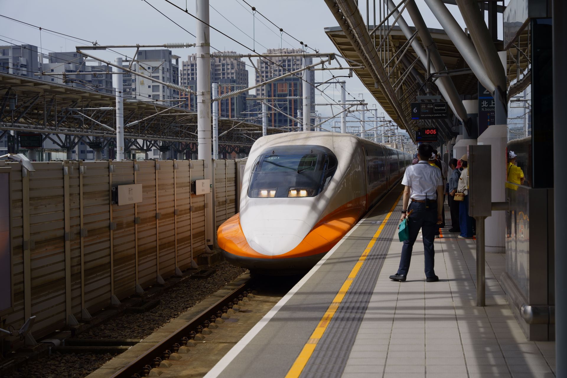 A white and orange Taiwan High Speed Rail train pulls into Hsinchu Station as a staff member and passengers wait on the platform